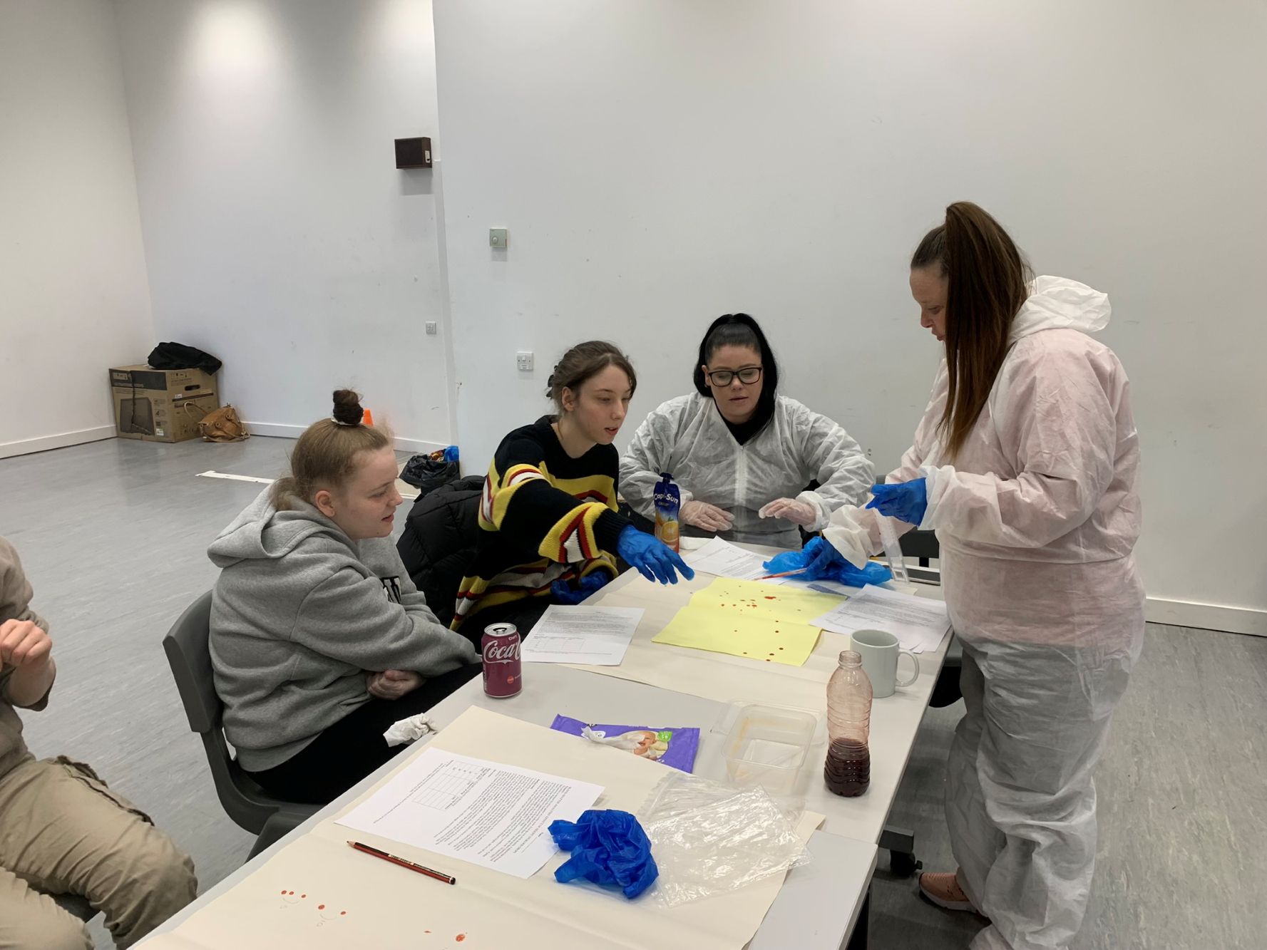 Several people seated around a table with papers, gloves, and bottles, engaged in an activity. One person in a pink protective suit stands at the end of the table, interacting with the group. The table has yellow sheets and scattered items related to the task.)
