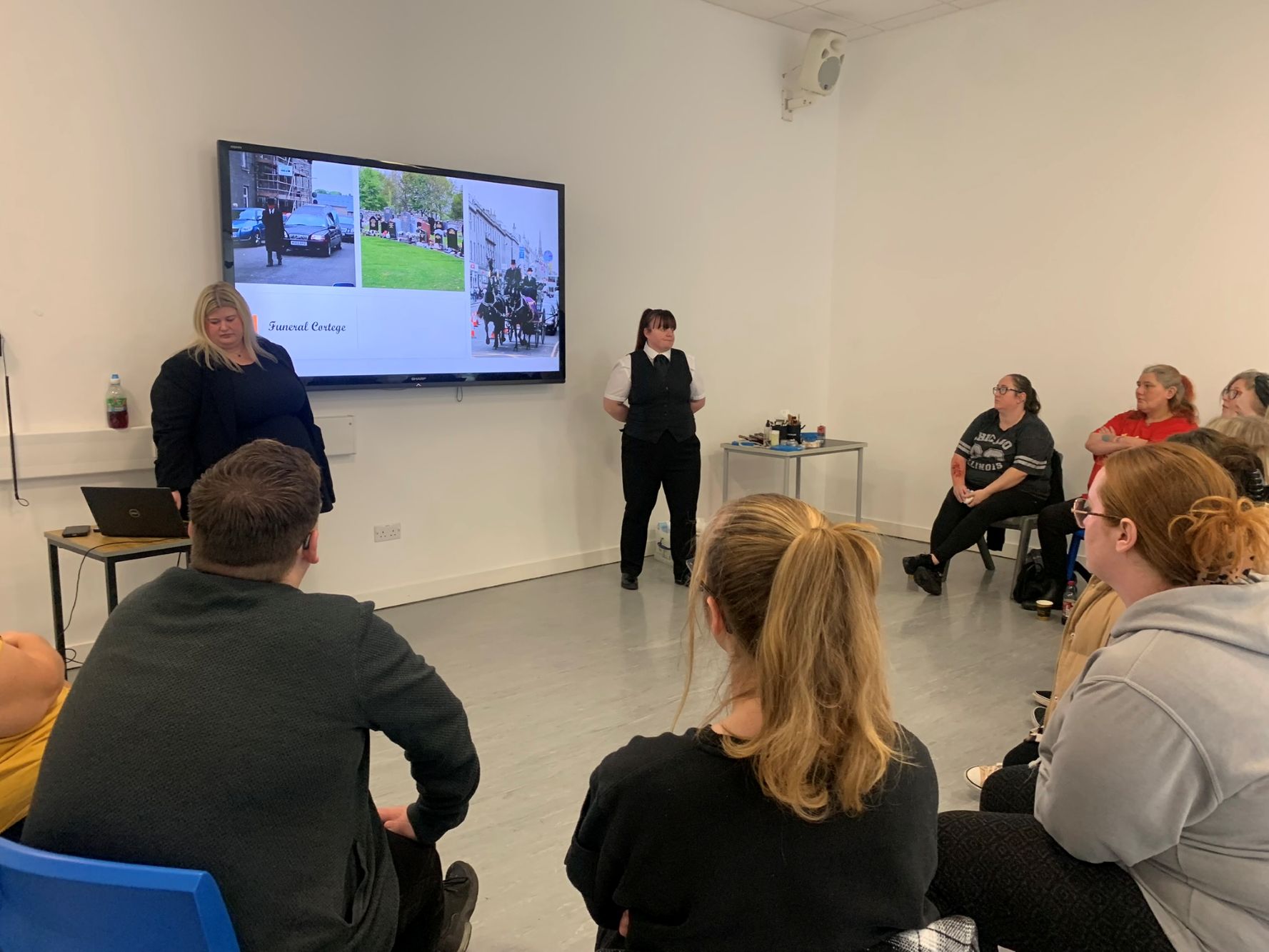 A classroom setting where a group of people is seated facing a large screen. The screen shows two images of outdoor scenes. Two presenters stand near the screen, one beside a laptop on a small table.