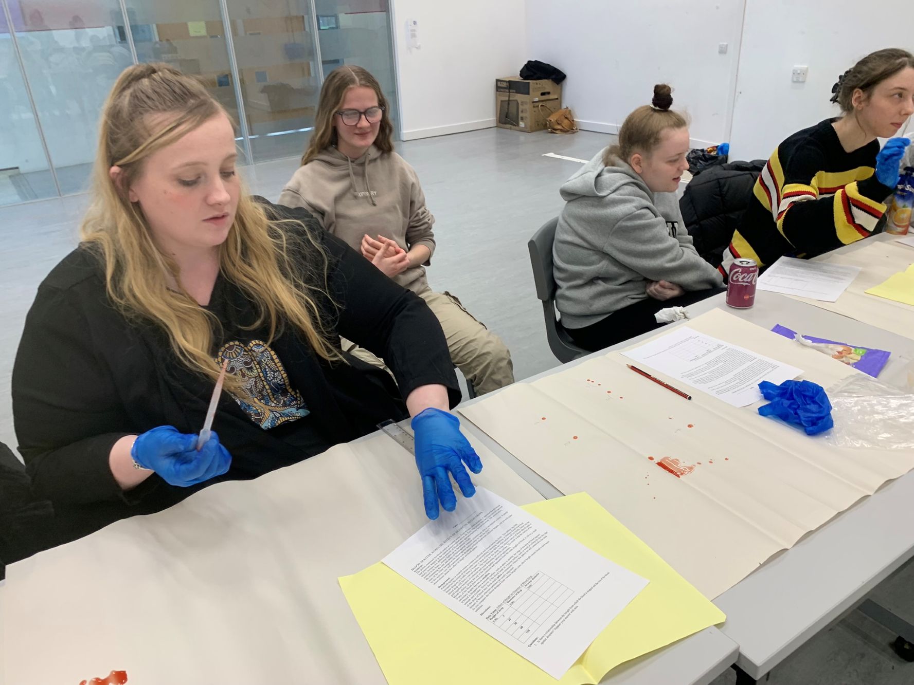 A group of people seated at a table with papers, gloves, and small containers. One person in the foreground is wearing blue gloves and holding a pipette. There are scattered red marks on the table, possibly from an experiment or demonstration.