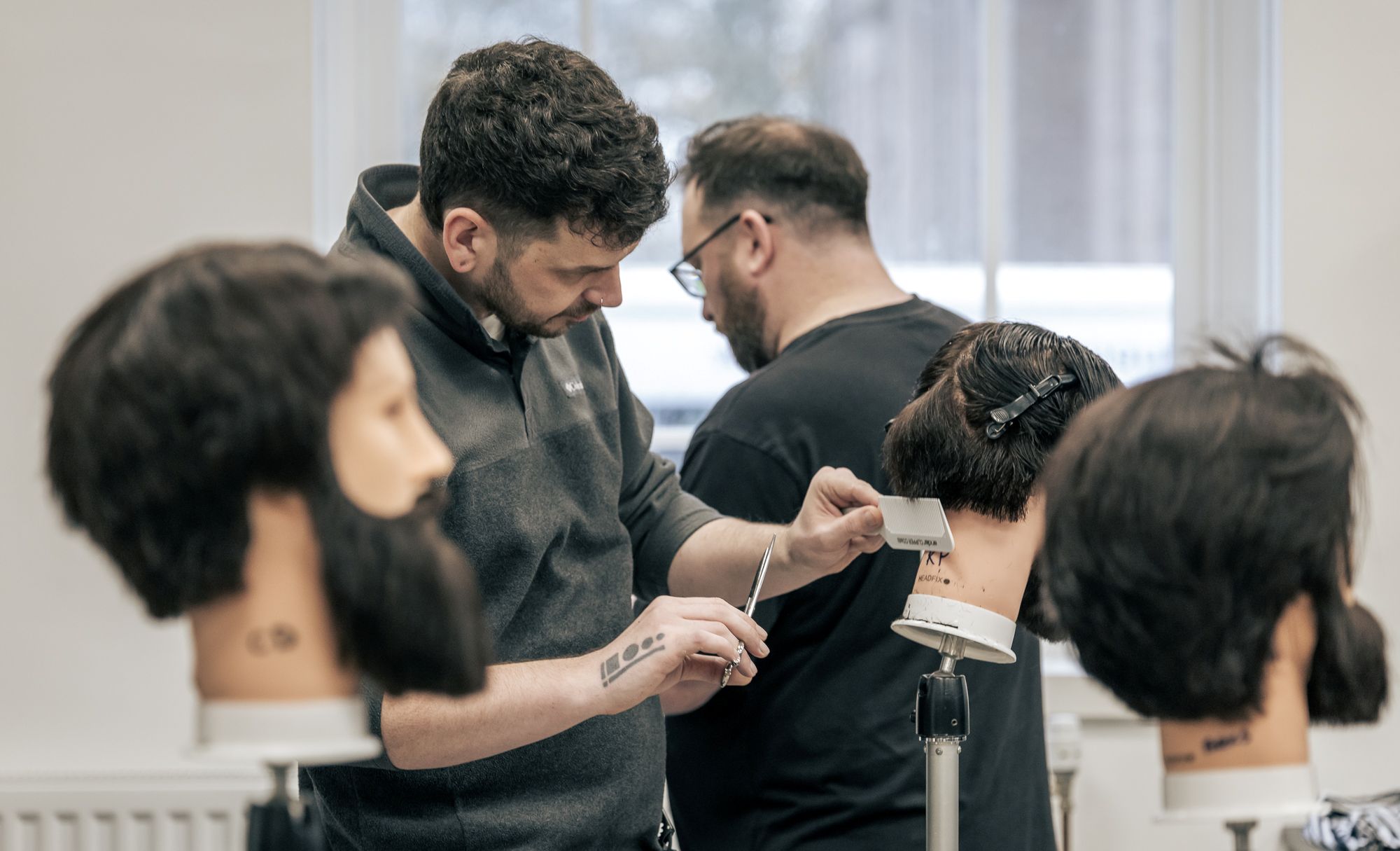 People practicing barbering techniques on mannequin heads in a classroom setting.