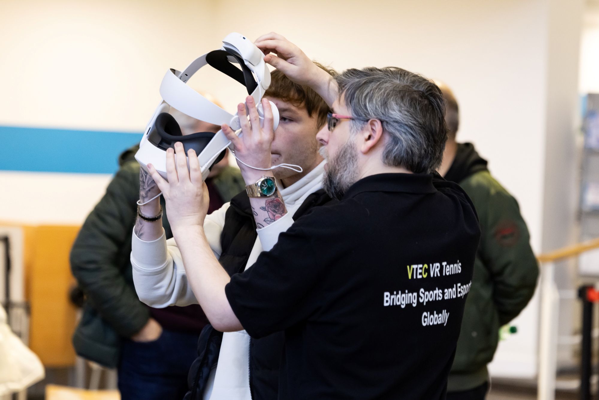 Individual assisting another with adjusting a VR headset, wearing a shirt with VTEC VR Tennis branding.