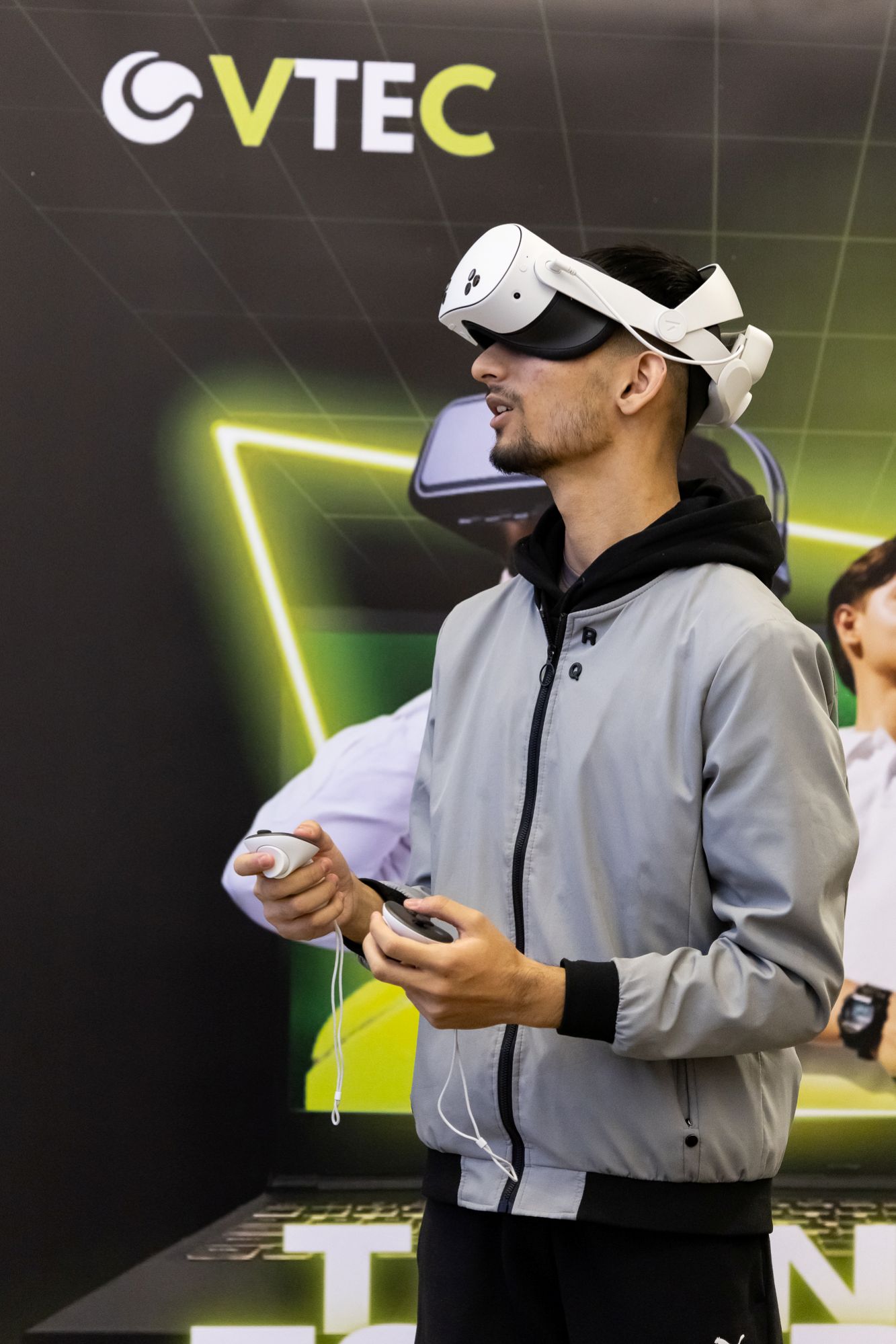 Person wearing a VR headset and holding controllers, standing in front of a VTEC promotional banner.