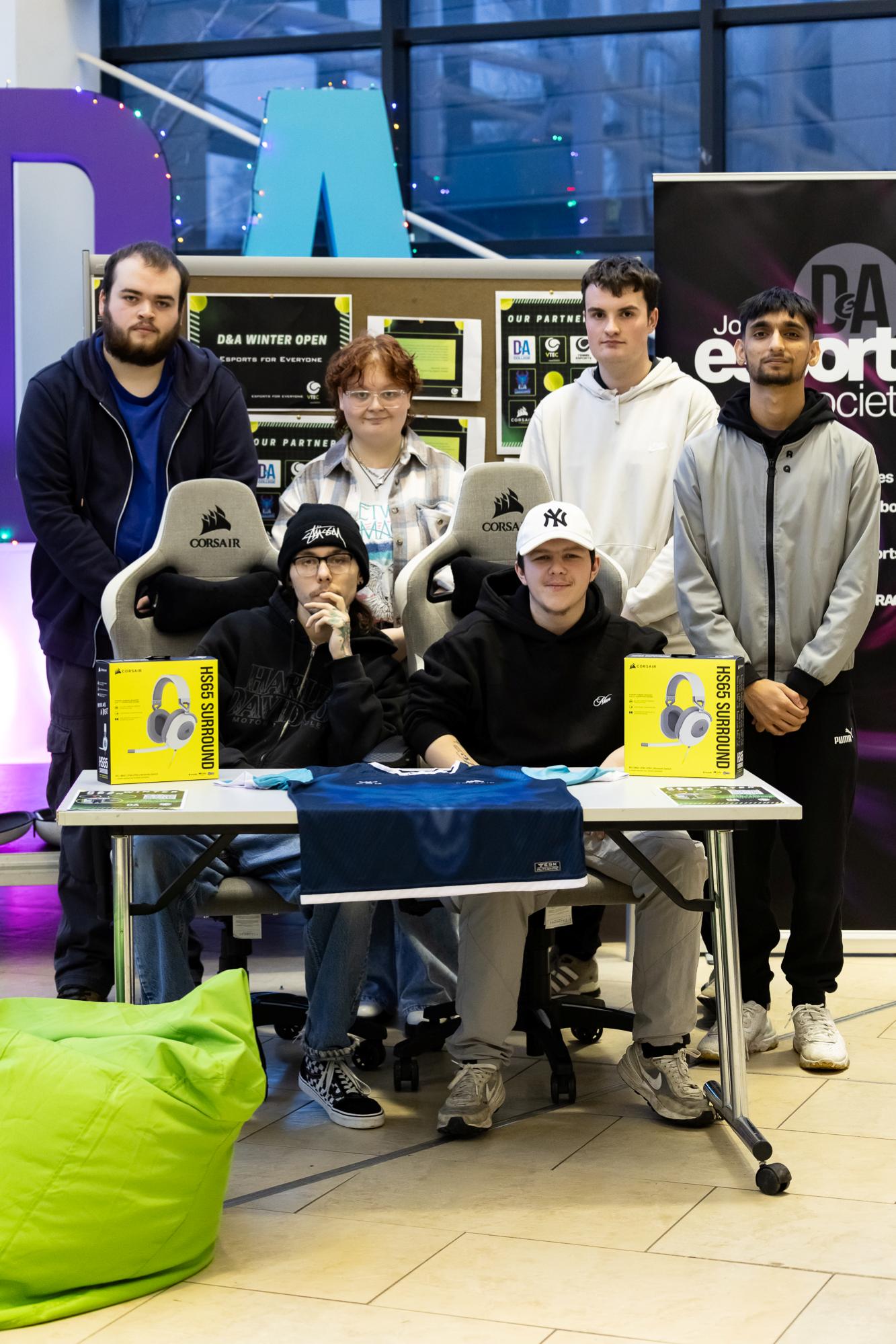 Group gathered around a gaming setup with branded chairs and Corsair headset boxes on the table.