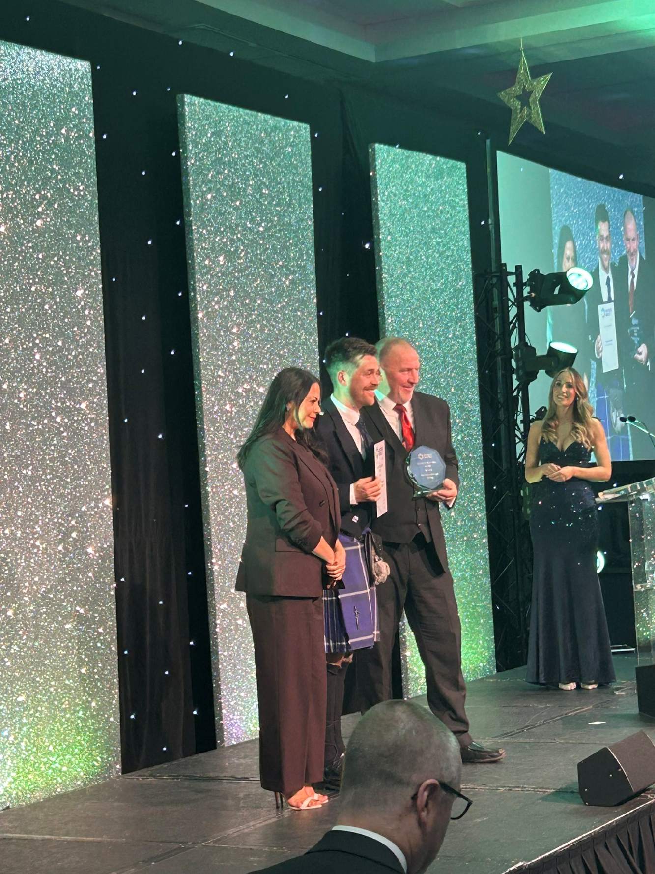 A group of people standing on a stage at an awards event, with a large branded backdrop displaying “CDN Awards.” Several individuals are holding trophies, and the stage is lit with bright spotlights.