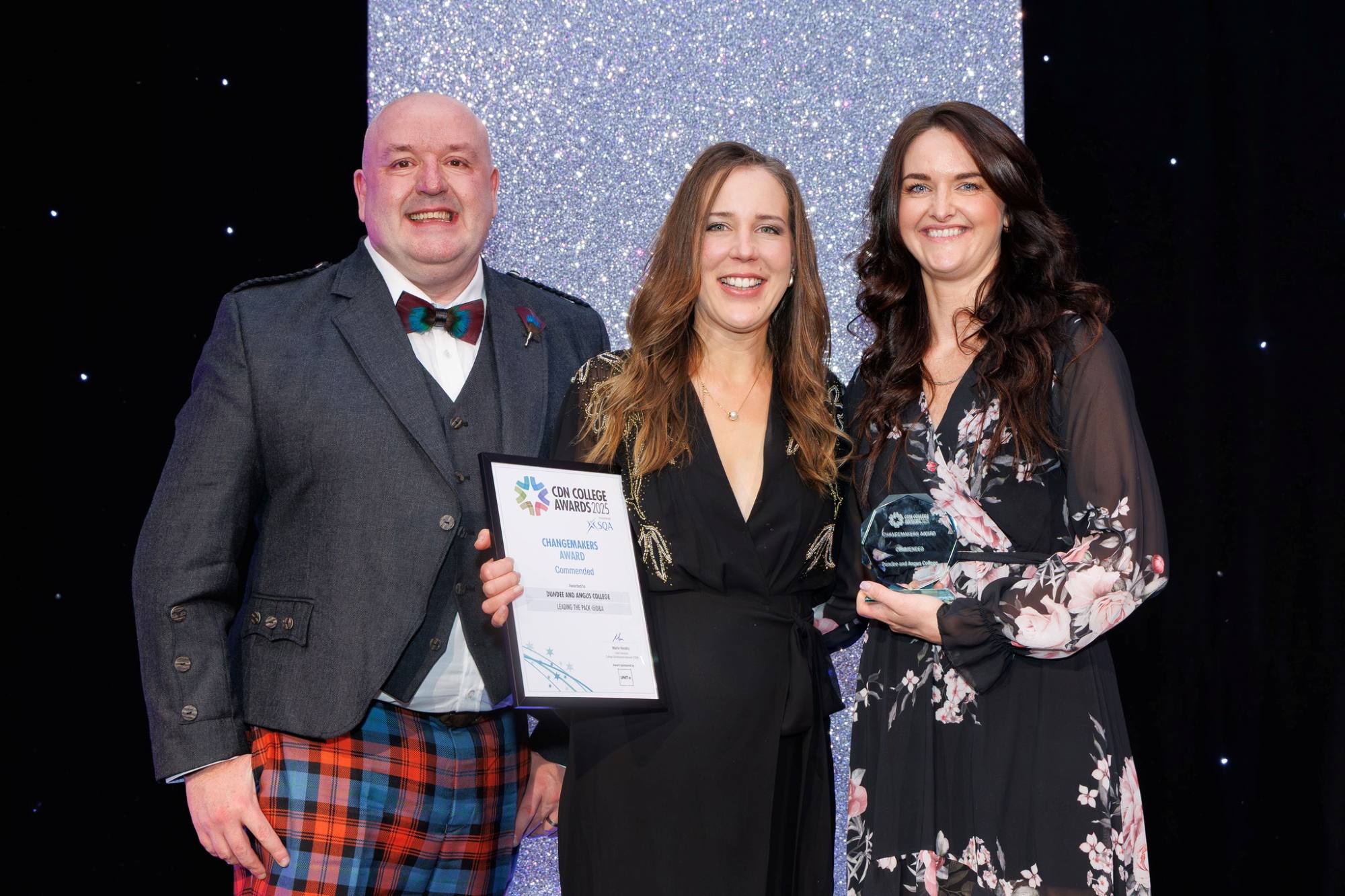 Three people against a glitter backdrop at an award ceremony. The middle person holds a certificate.