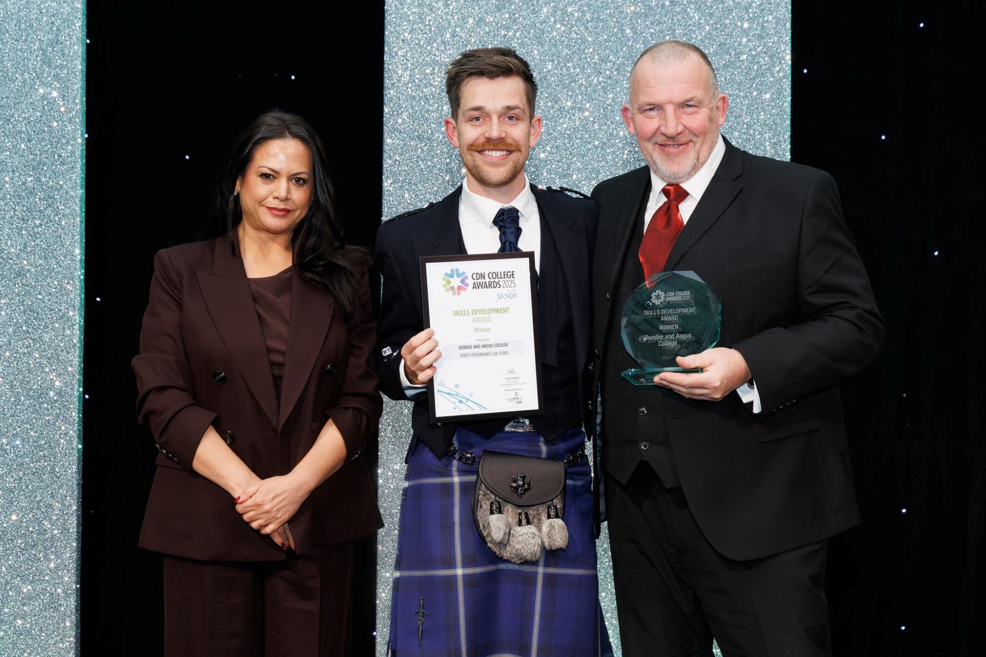Three people against a glitter backdrop at an award ceremony. The middle person holds a certificate and the right person holds a glass trophy.)