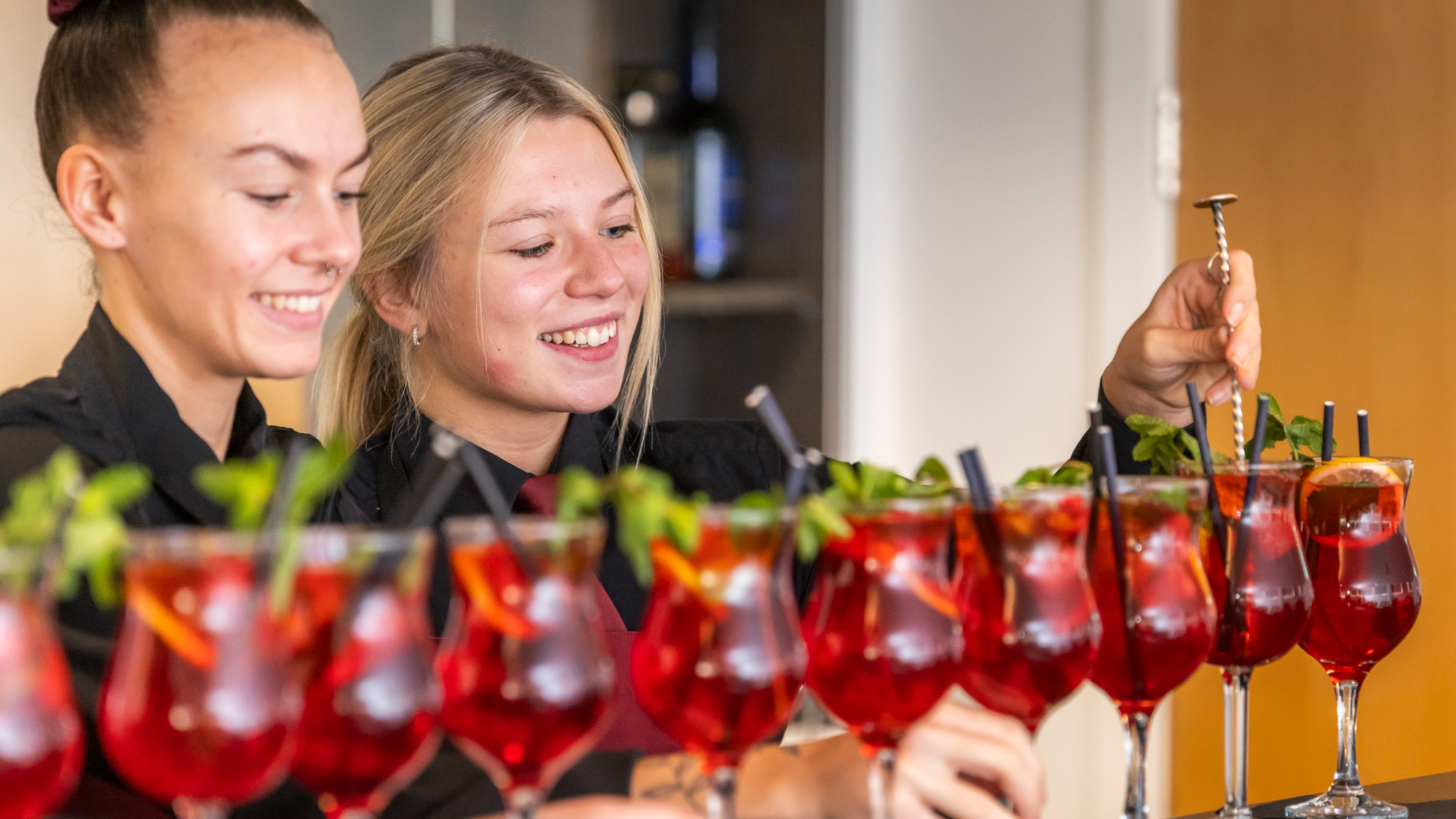 Two hospitality students in black shirts stir a row of red cocktails in glasses.