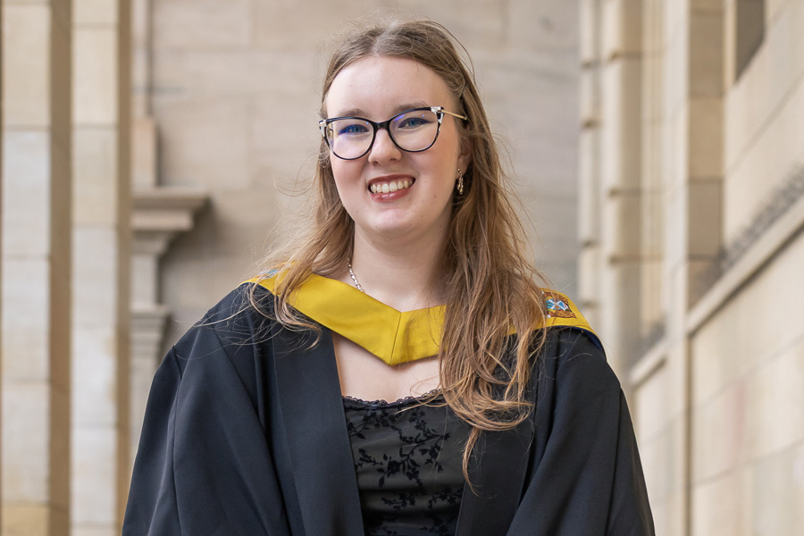 Student standing outside of Caird Hall smiling wearing graduation robes