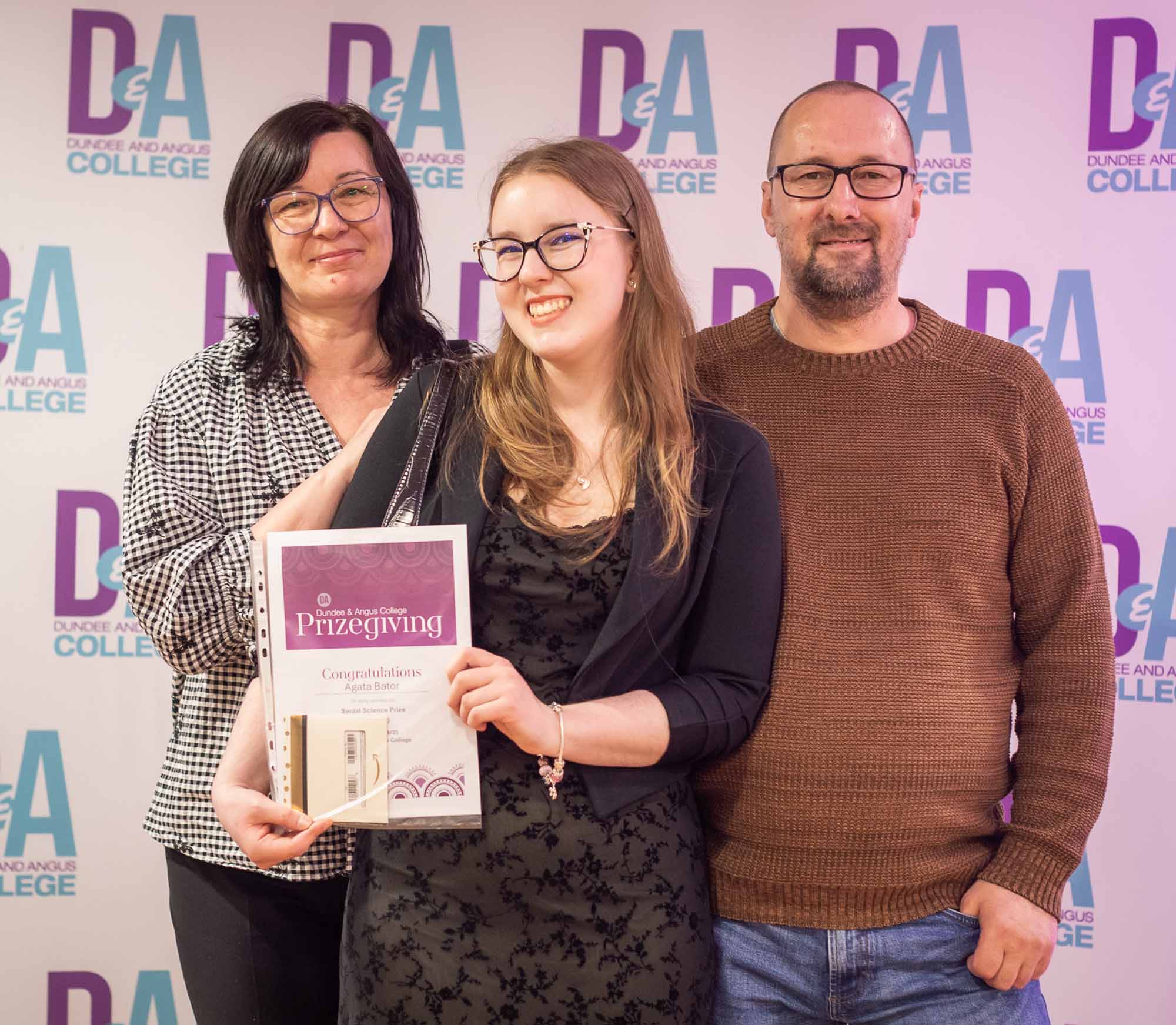 Student standing in the middle of parents holding certificate and prize in front of Dundee and Angus College backdrop)