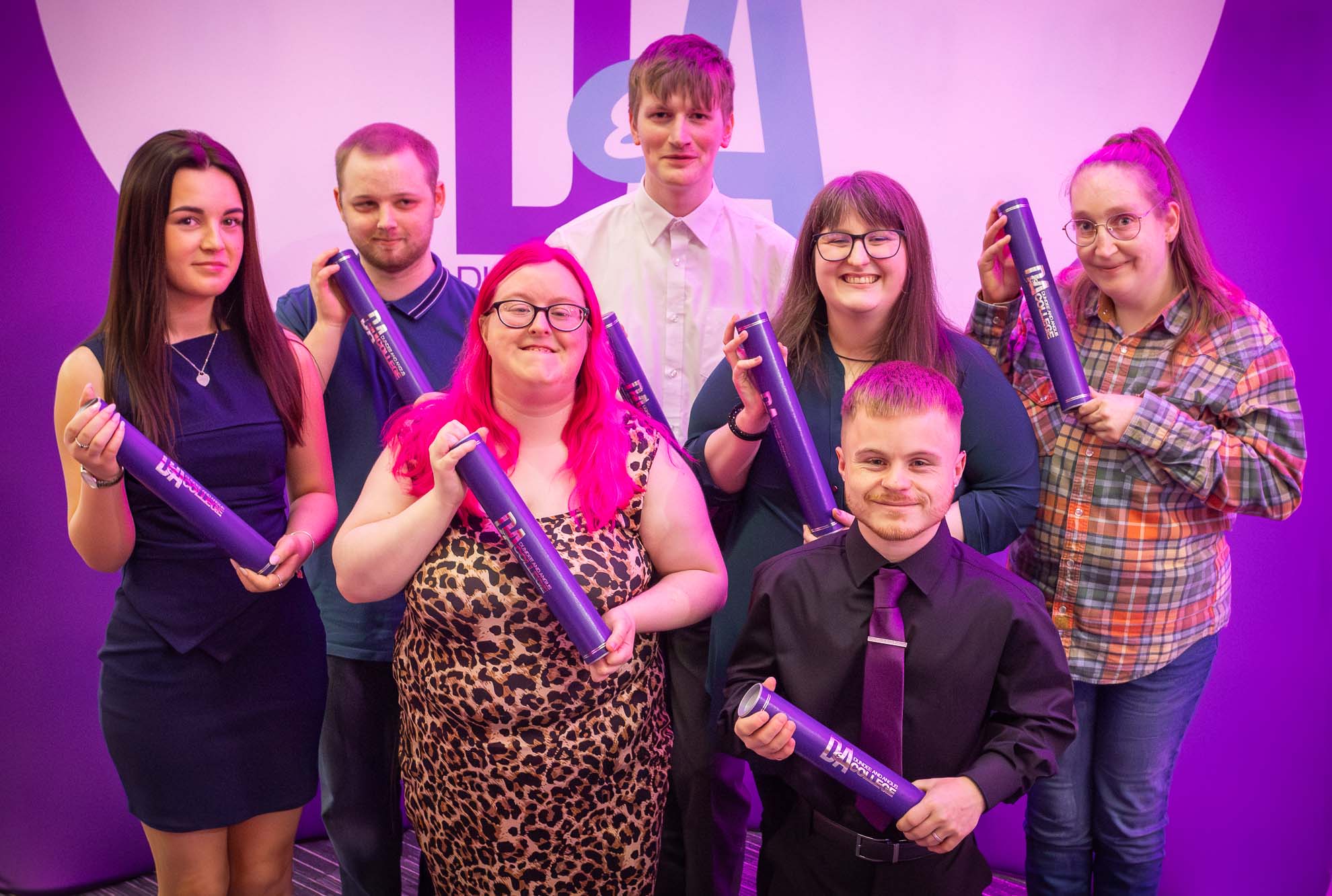 A group of seven students standing together, each holding a purple certificate tube. A large purple and white banner with the letters 'DA' is visible in the background. The individuals' faces are blurred, and they are dressed in a mix of formal and casual clothing.)