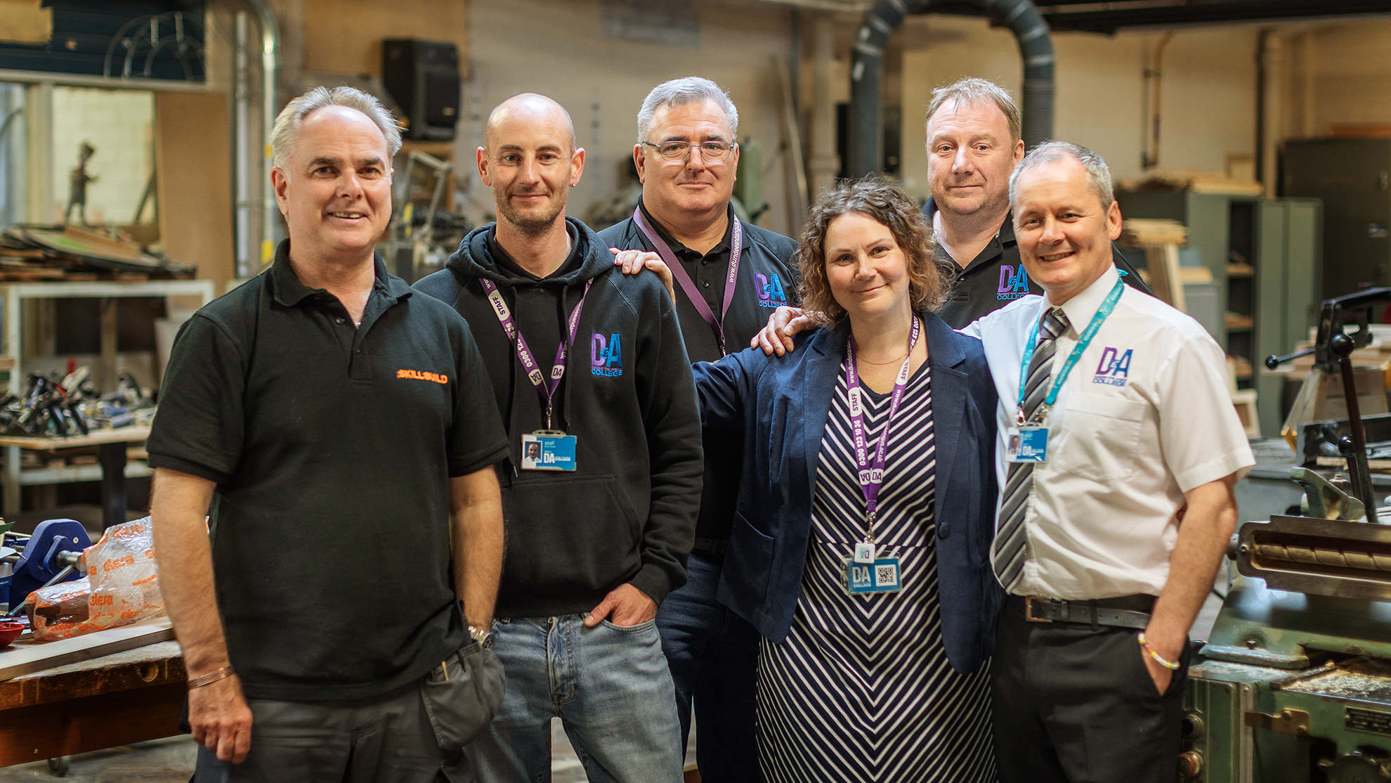 A group of six people standing together in a workshop or industrial setting. They are wearing various types of clothing, including polo shirts, hoodies, and a dress with a blazer. Each person has an identification badge on a lanyard around their neck. The background shows workbenches, tools, and machinery typically found in a workshop environment)