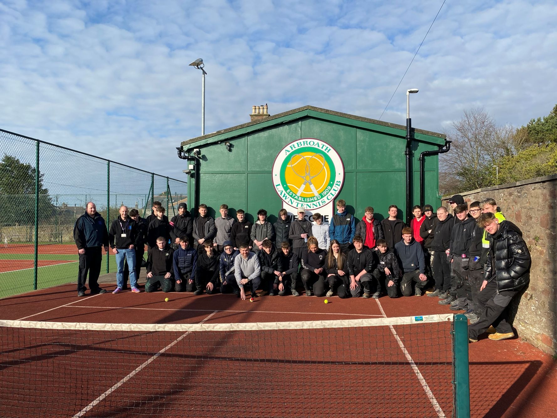 Group photo of around 40 people on a red clay tennis court in front of a green building labeled 'Arbroath Lawn Tennis Club.' Some individuals are standing while others are crouching, all in casual attire. A tennis net is visible in the foreground under a partly cloudy sky)